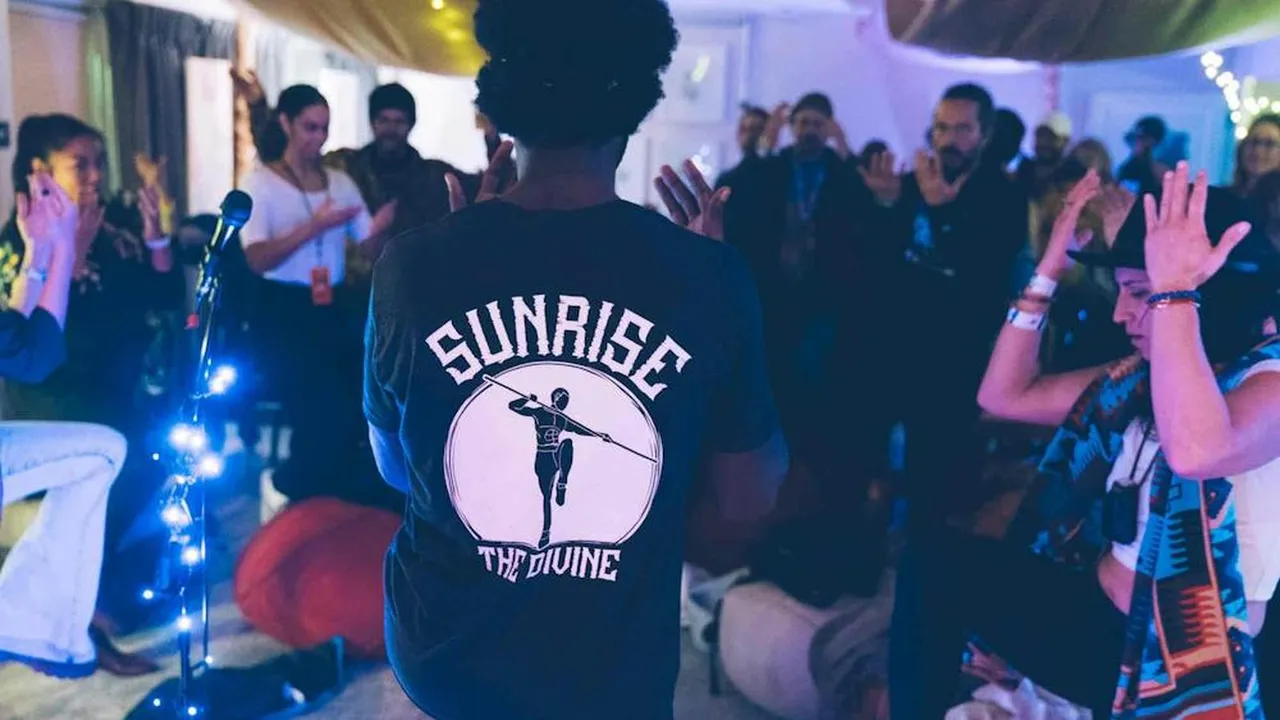 Sunrise The Divine, seen from behind in a Sunrise The Divine shirt, leading an indoor Trap Chi session as the group raises their hands.