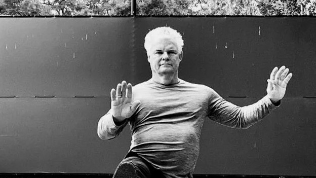 Instructor Skee Goedhart in a tai chi posture with both palms raised on an outdoor court with trees behind the fence, black and white photo