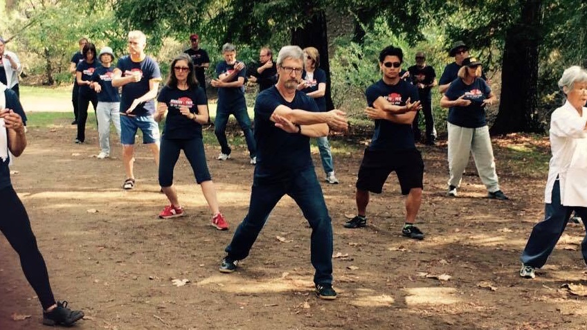 More than a dozen people, most in matching navy T-shirts, practicing tai chi together on a shaded dirt clearing under tall trees