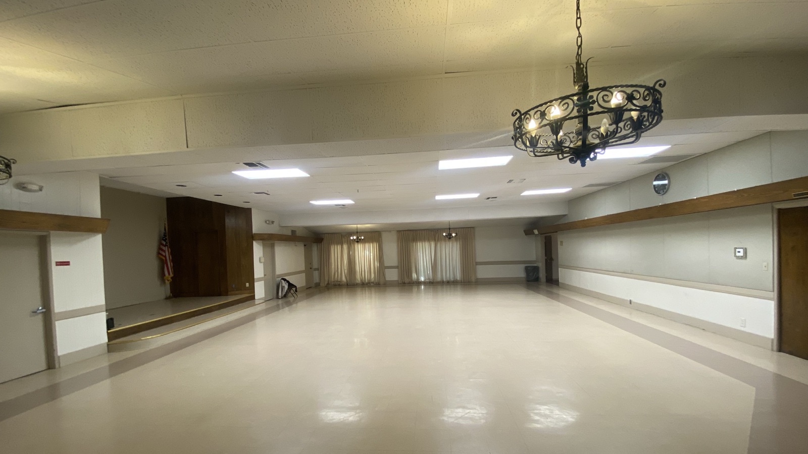 An empty hall at the Encino Women's Club in the Encino Community Center, with a small stage and a flag, an iron chandelier, curtained windows at the far end and a wide open floor