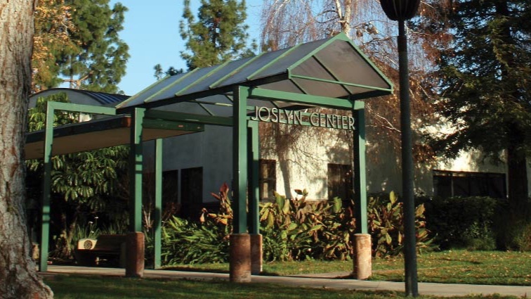 A green covered walkway with the Joslyn Center sign leads to the building in Burbank, framed by tall trees, a lamp post and garden plantings.