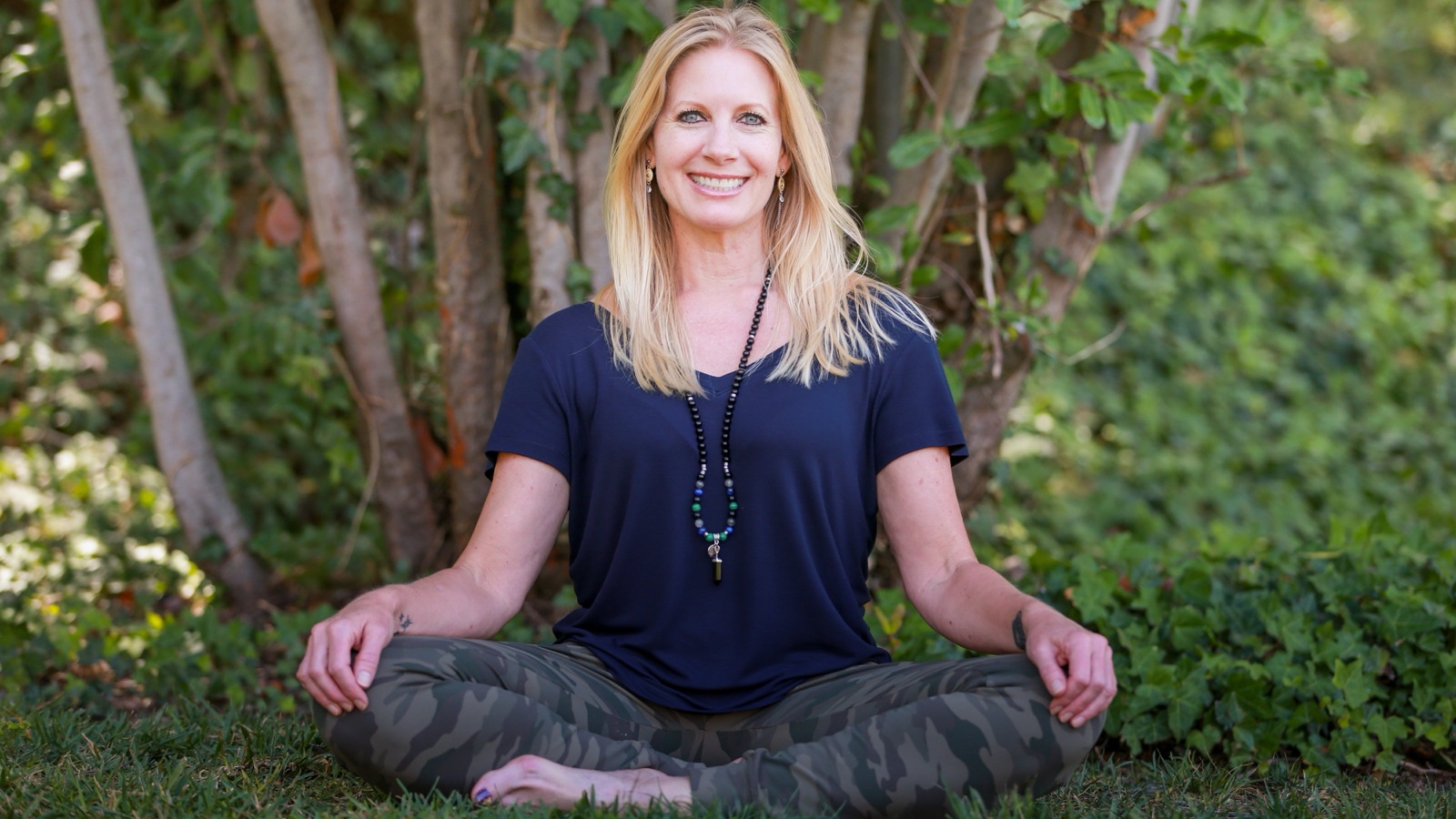 Treeesa Nieto sits cross-legged on the grass in front of leafy trees, hands resting on her knees, smiling at the camera