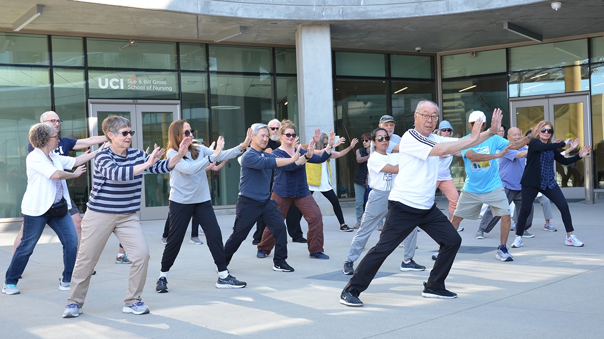 A teacher in a white shirt leads a large group through a tai chi form on the outdoor plaza in front of the UCI Sue & Bill Gross School of Nursing building